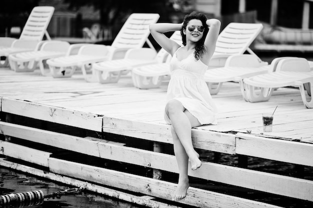 Retrato de uma jovem fantástica com óculos de sol e sua bebida sentada no porto de madeira branco ao lado da água foto em preto e branco