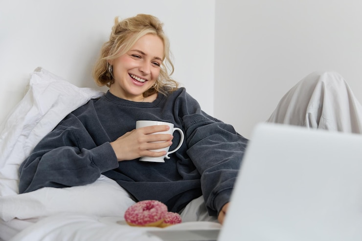 Foto grátis retrato de uma jovem feliz deitada na cama olhando para a tela do laptop segurando uma chávena de chá e