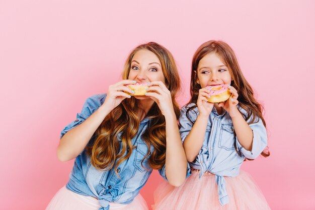 Retrato de uma jovem feliz mãe e filha comendo rosquinhas saborosas depois do jantar, isolado no fundo rosa. Duas adoráveis irmãs encaracoladas de cabelos compridos assaram deliciosos bolinhos juntas e os provaram.