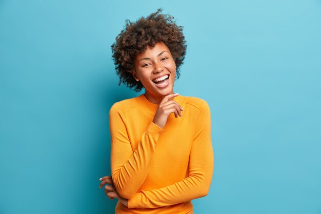 Retrato de uma jovem feliz rindo, segurando a mão no queixo, expressa emoções positivas, sorri amplamente, tem uma expressão despreocupada, usa um macacão laranja isolado sobre a parede azul