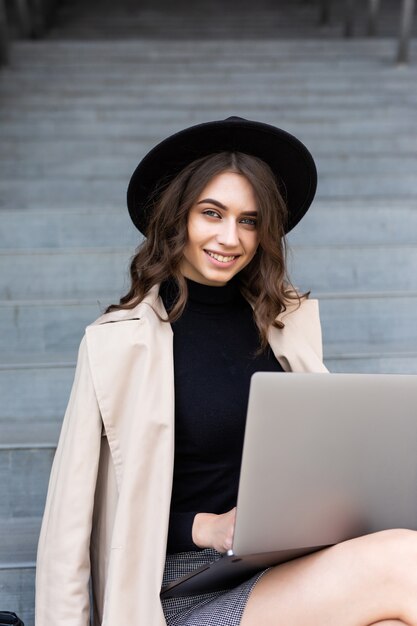 Retrato de uma jovem feliz sentada na escada da cidade usando um laptop ao ar livre