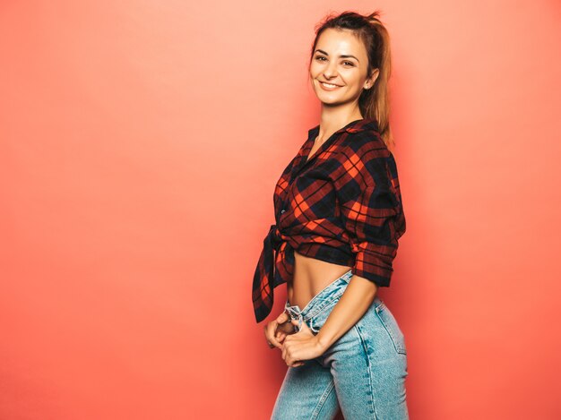 Retrato de uma jovem garota bonita sorridente hipster em roupas de verão jeans e camisa quadriculada na moda. Mulher despreocupada "sexy" que levanta perto da parede cor-de-rosa no estúdio. Modelo positivo sem maquiagem