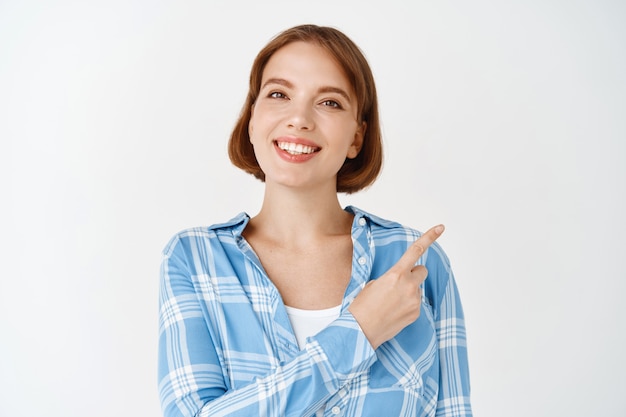 Retrato de uma jovem garota linda em uma camisa, apontando para o canto superior direito, sorrindo. mulher com cabelo curto e sorriso branco faz anúncio