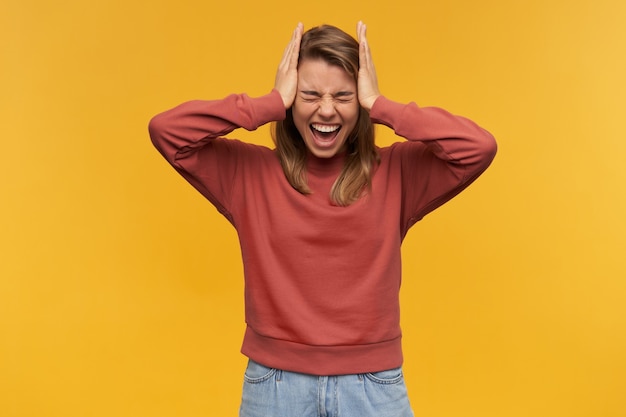 Foto grátis retrato de uma jovem gritando com raiva em um moletom de terracota com os olhos fechados e as mãos na cabeça, gritando e tendo uma dor de cabeça isolada na parede amarela