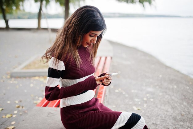 Retrato de uma jovem linda indiana ou adolescente do sul da Ásia em vestido sentado no banco com telefone celular