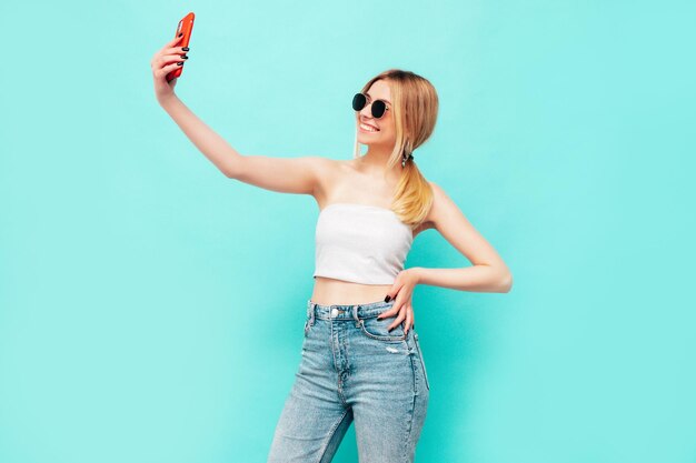 Retrato de uma jovem linda loira sorridente em roupas da moda de verão Mulher despreocupada sexy posando perto da parede azul no estúdio Modelo positivo se divertindo dentro de casa Alegre e feliz tomando selfie