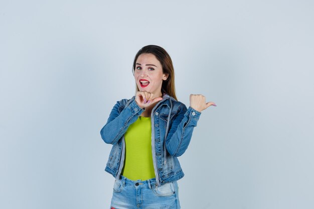 Retrato de uma jovem linda mulher apontando para a direita com os polegares em roupa jeans e olhando alegre para a frente