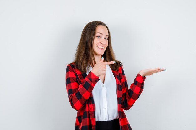 Retrato de uma jovem linda mulher apontando para a direita, espalhando a palma da mão para o lado em uma roupa casual e olhando para a frente alegre