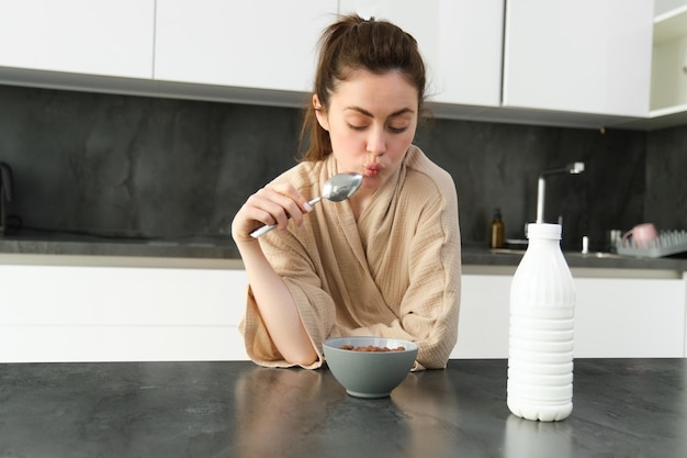 Foto grátis retrato de uma jovem linda mulher de roupão comendo cereais no café da manhã apoia-se na bancada da cozinha