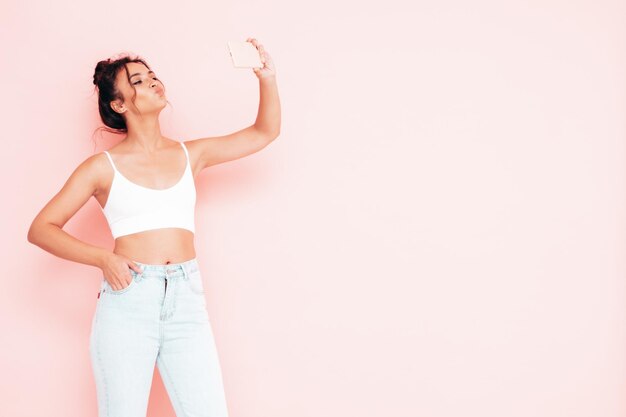 Retrato de uma jovem linda mulher sorridente em roupas da moda de verão Mulher despreocupada sexy posando perto da parede rosa no estúdio Modelo positivo se divertindo dentro de casa Tirando fotos de selfie