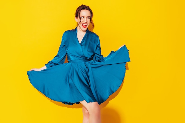 Retrato de uma jovem linda mulher sorridente no vestido azul de verão na moda Mulher despreocupada sexy posando perto da parede amarela no estúdio Modelo positivo se divertindo e enlouquecendo Alegre e feliz