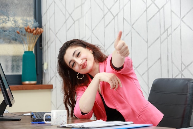 Foto grátis retrato de uma jovem linda sentada no escritório e gesticulando o polegar para cima foto de alta qualidade