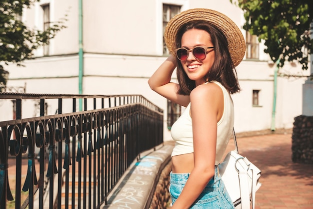 Retrato de uma jovem linda sorridente hipster em shorts jeans de verão na moda e chapéu Modelo despreocupado sexy posando no fundo da rua ao pôr do sol Modelo positivo ao ar livre com maleta