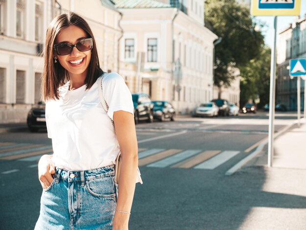 Retrato de uma jovem linda sorridente hipster em shorts jeans na moda verão Modelo despreocupado sexy posando no fundo da rua ao pôr do sol Modelo positivo ao ar livre em óculos de sol