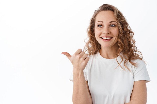 Retrato de uma jovem loira apontando olhando para a esquerda sorrindo satisfeita ao verificar uma boa oferta promocional sobre fundo branco