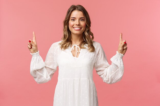 Retrato de uma jovem loira bonita confiante em um vestido branco bonito, apontando os dedos para cima no anúncio superior, olhando para a câmera com um sorriso radiante, parede rosa de pé.