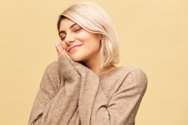 Retrato de uma jovem loira cansada com sono, com argola no nariz, colocando a cabeça nas mãos pressionadas juntas e mantendo os olhos fechados, tirando uma soneca ou dormindo, sorrindo alegremente. conceito de sono, roupa de cama e fadiga