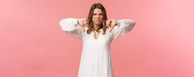 Retrato de uma jovem loira caucasiana exigente de vestido branco sobre fundo rosa expressando desagrado