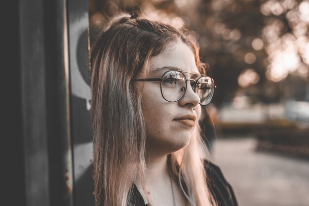 Retrato de uma jovem loira com óculos encostado em uma parede e sorrindo gentilmente