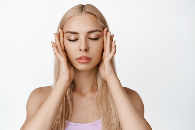 Retrato de uma jovem loira com pele perfeita brilhante, feche os olhos e toque a pele facial macia e saudável, aplicando toner sobre fundo branco