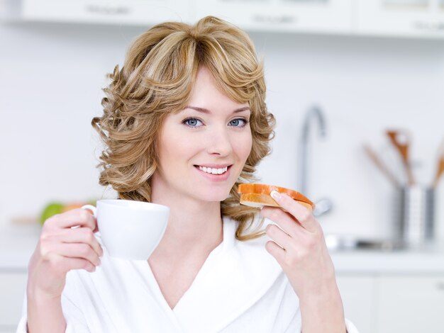 Retrato de uma jovem loira com um sorriso feliz tomando um café da manhã na cozinha