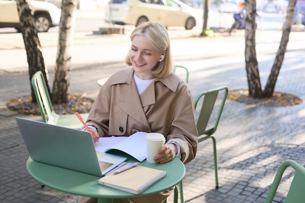 Foto grátis retrato de uma jovem loira estudante em um café de rua usando fones de ouvido sem fio usando um laptop