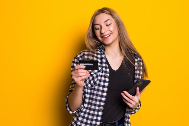 Retrato de uma jovem loira feliz, mostrando um cartão de crédito de plástico ao usar um telefone celular isolado na parede amarela