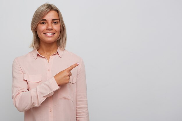 Foto grátis retrato de uma jovem loira linda e alegre com aparelho nos dentes e usa uma camisa rosa parece confiante e aponta para o lado com o dedo isolado sobre a parede branca
