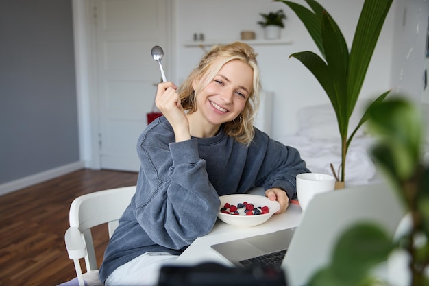 Retrato de uma jovem loira sorridente comendo na frente do laptop vendo vídeos online enquanto tem
