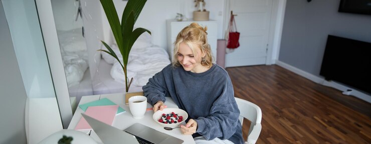 Foto grátis retrato de uma jovem loira sorridente comendo na frente do laptop vendo vídeos online enquanto tem
