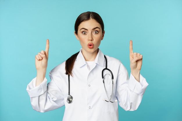 Retrato de uma jovem médica surpresa, trabalhadora médica apontando os dedos para cima ofegante, mostrando ...