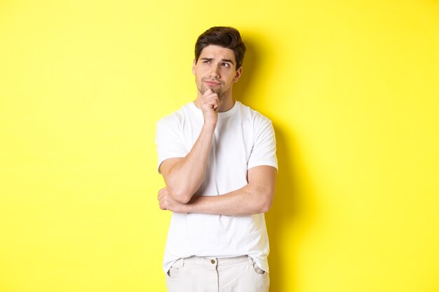 Retrato de uma jovem modelo masculino pensando, olhando para o canto superior esquerdo e fazendo a escolha, em pé perto do espaço da cópia, fundo amarelo.