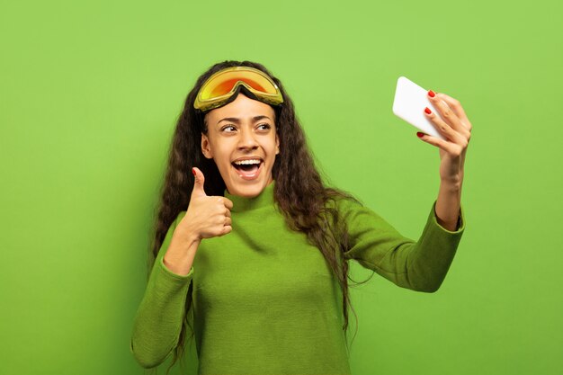 Retrato de uma jovem morena afro-americana na máscara de esqui no fundo verde do estúdio. Conceito de emoções humanas, expressão facial, vendas, anúncio, esporte de inverno e feriados. Fazendo selfie ou vlog.