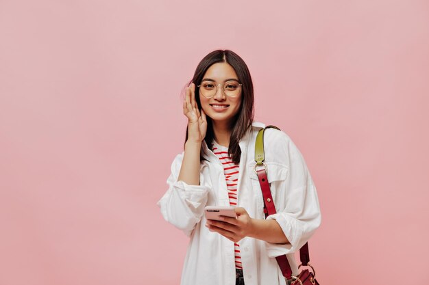 Retrato de uma jovem morena bronzeada em óculos e camisa branca segura celular e sorri em fundo isolado rosa