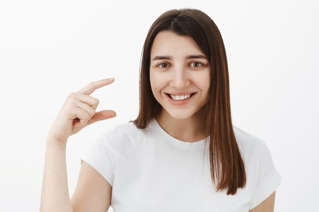 Retrato de uma jovem morena feliz, simpática e otimista, simpática e otimista em uma camiseta branca, sorrindo satisfeita ao modelar um pequeno ou minúsculo objeto na mão, falando sobre um pequeno produto sobre a parede cinza