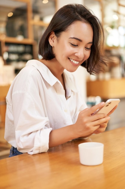 Retrato de uma jovem morena sentada com café e usando smartphone em um café conversando no mobi