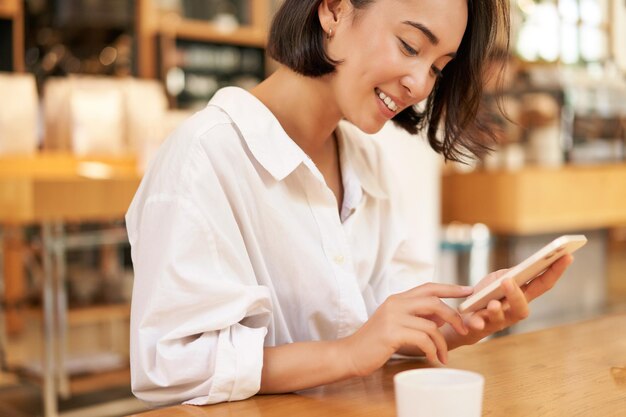 Retrato de uma jovem morena sentada com café e usando smartphone em um café conversando