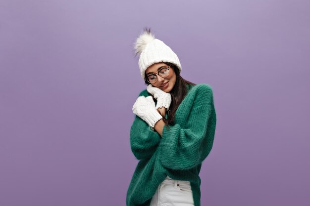 Retrato de uma jovem morena tímida de chapéu de malha branca e luvas Garota de cabelos compridos em óculos e poses de suéter verde sobre fundo roxo