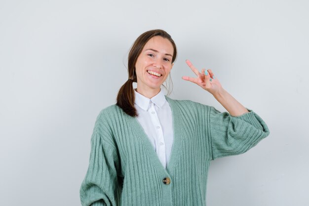 Retrato de uma jovem mostrando o V-sign de blusa, casaco de lã e olhando alegre para a frente