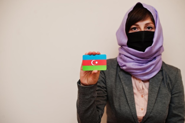 Retrato de uma jovem muçulmana vestindo roupa formal protege a máscara facial e o lenço de cabeça hijab segura o cartão da bandeira do azerbaijão contra o conceito de país coronavirus isolado de fundo