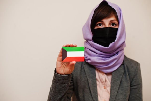 Foto grátis retrato de uma jovem muçulmana vestindo roupa formal protege a máscara facial e o lenço de cabeça hijab segura o cartão da bandeira do kuwait contra o conceito de país coronavirus isolado de fundo