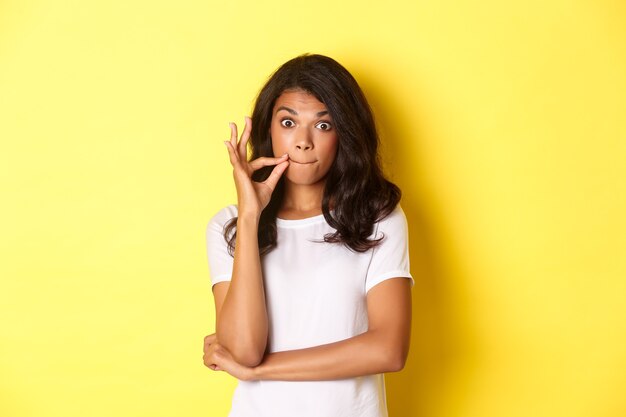 Retrato de uma jovem mulher afro-americana, fazendo promessa de guardar segredo, selar os lábios, fechando a boca com os dedos, em pé sobre um fundo amarelo.