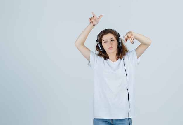 Retrato de uma jovem mulher apontando para longe enquanto ouve música com fones de ouvido em uma camiseta branca, jeans e uma vista frontal de um sonho