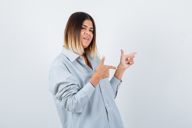 Retrato de uma jovem mulher apontando para o lado direito com uma camisa grande e olhando frontalmente feliz