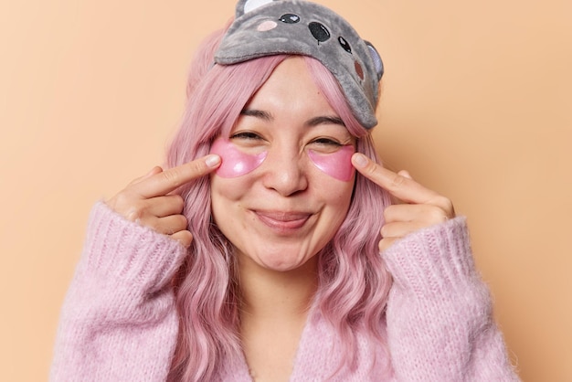 Retrato de uma jovem mulher asiática com cabelo longo tingido de rosa aponta para manchas de hidrogel sob os olhos se sente bem submetido a tratamentos de beleza usa máscara de dormir e jumper isolado sobre fundo bege.