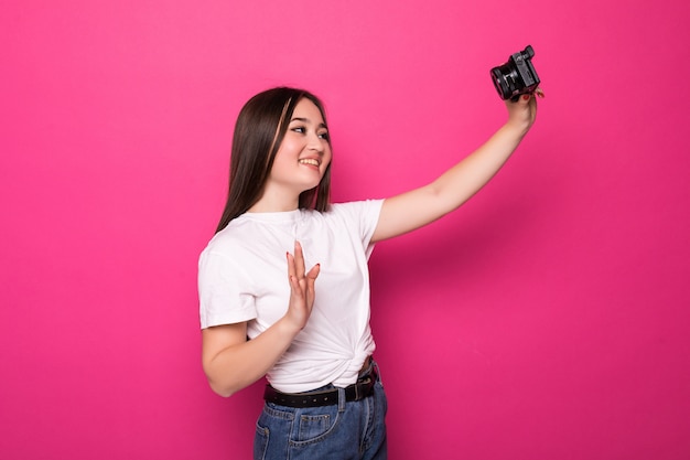 Retrato de uma jovem mulher asiática feliz vestida de vestido branco e chapéu de verão segurando a câmera fotográfica e olhando para o espaço da cópia sobre parede rosa