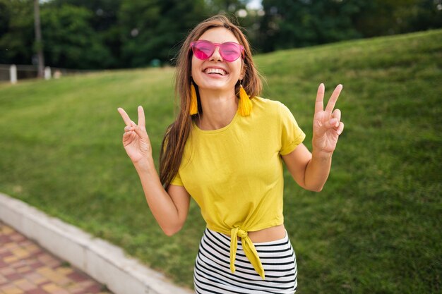 Retrato de uma jovem mulher atraente e elegante, posando no parque da cidade, sorrindo, alegre, positiva, vestindo blusa amarela, minissaia listrada, bolsa, óculos de sol rosa, tendência da moda no estilo de verão