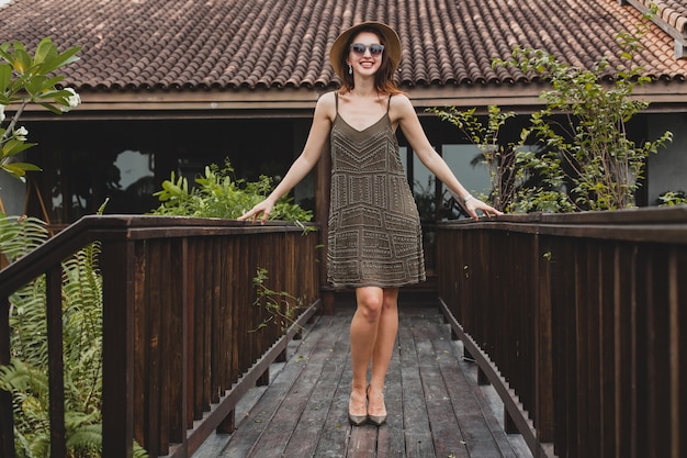 Foto grátis retrato de uma jovem mulher atraente em um vestido elegante, chapéu de palha, estilo de verão, tendência da moda, férias, sorrindo, acessórios elegantes, óculos de sol, posando em villa tropical em bali