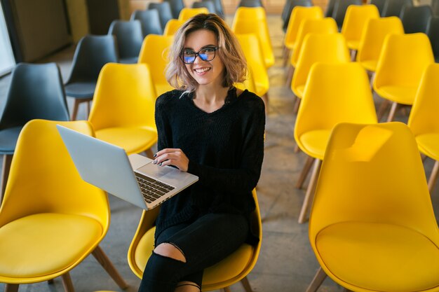 Retrato de uma jovem mulher atraente, sentado na sala de aula, trabalhando no laptop usando óculos, aluno aprendendo na sala de aula com muitas cadeiras amarelas