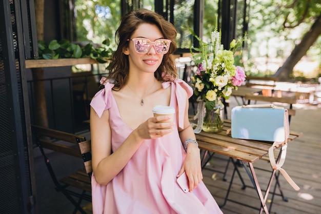 Retrato de uma jovem mulher atraente, sentado no café com roupa de moda de verão, vestido de algodão rosa, óculos de sol, sorrindo, bebendo café, acessórios elegantes, relaxante, vestuário da moda